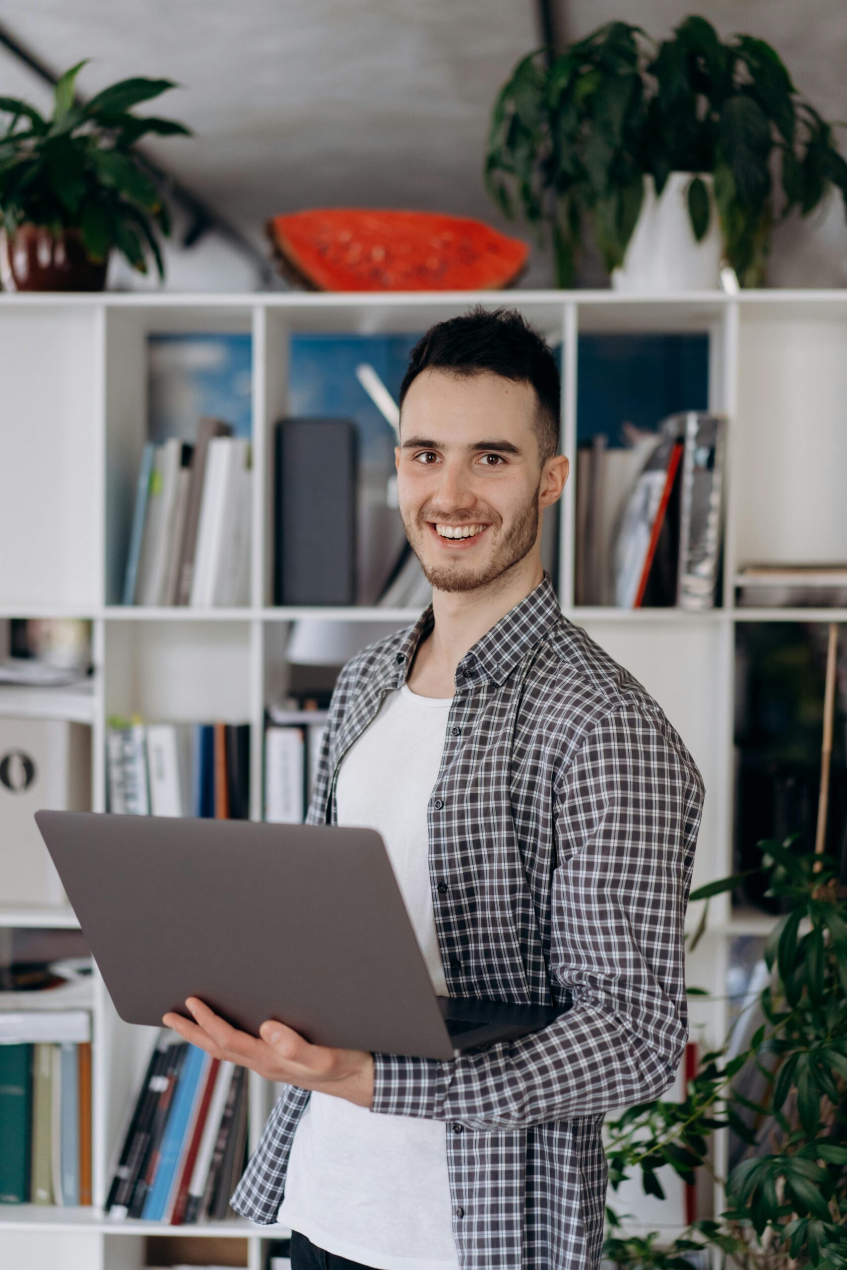 Young man in a casual checkered shirt holding a laptop in a modern indoor setting.